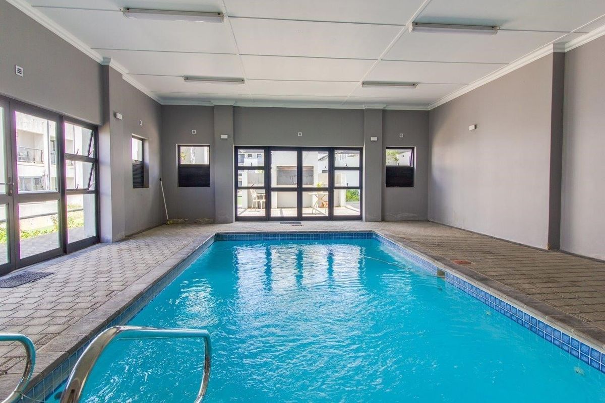 A well-maintained indoor swimming pool area featuring tiled surrounds, large aluminium-framed glass doors, and a clean, neutral interior finish.