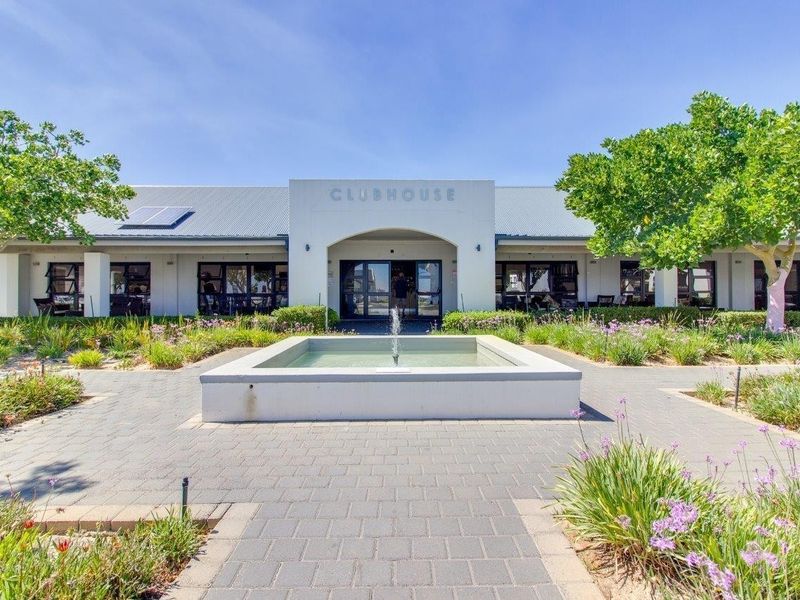 A well-maintained retirement village clubhouse featuring a central fountain, paved entrance walkway, and landscaped gardens under a clear blue sky.