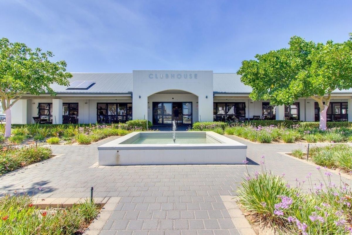 A well-maintained retirement village clubhouse featuring a central fountain, paved entrance walkway, and landscaped gardens under a clear blue sky.