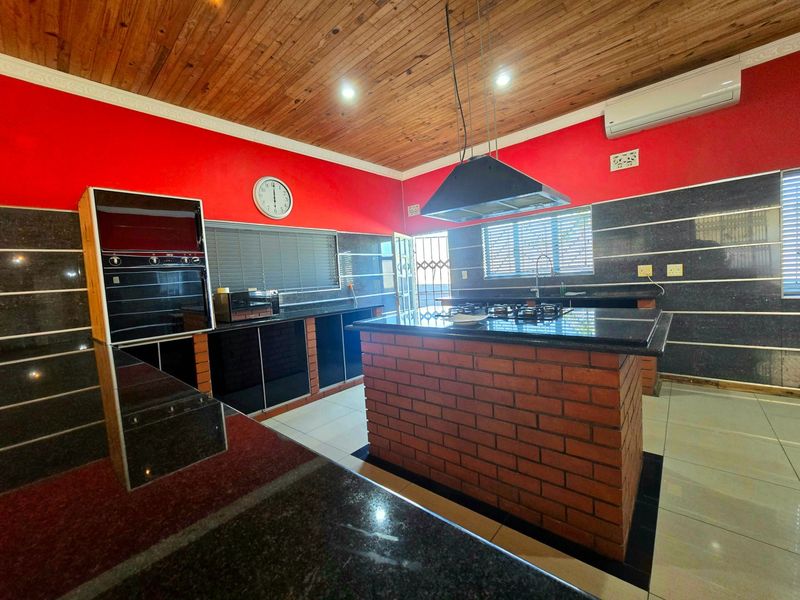 Spacious kitchen featuring granite countertops, a central brick island with a gas hob, built-in oven, and a distinctive timber ceiling.