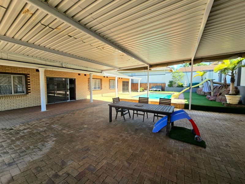 Spacious covered patio area featuring brick paving, overlooking a swimming pool and garden, providing an ideal space for outdoor entertainment.
