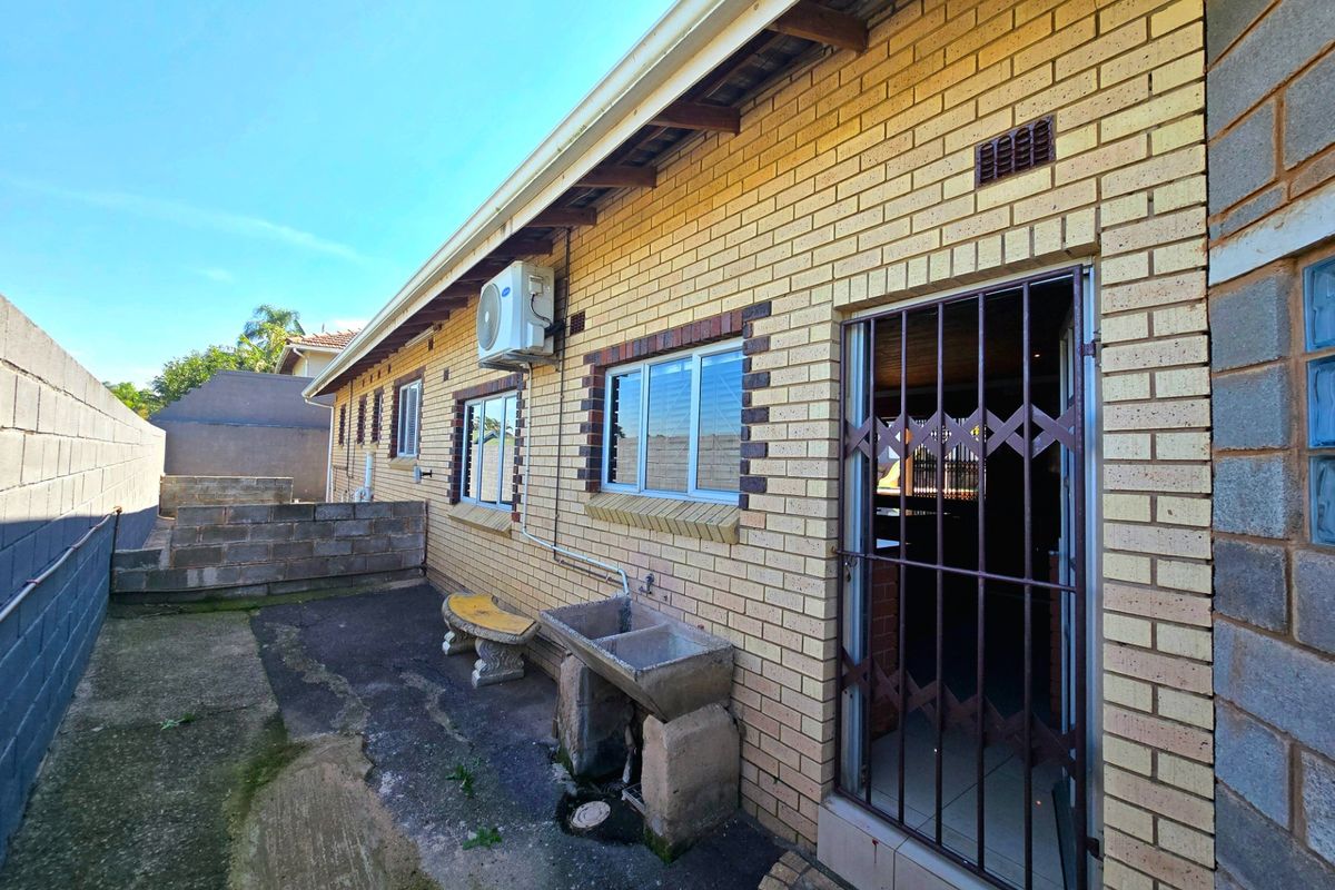 Rear exterior view of the house featuring a brick facade, security gate, wall-mounted air conditioning unit, and a concrete wash basin on a paved area.