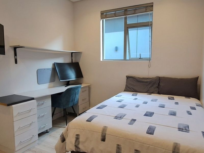 A compact bedroom featuring a built-in desk and shelving unit, neutral walls, and a window with a fitted blind.