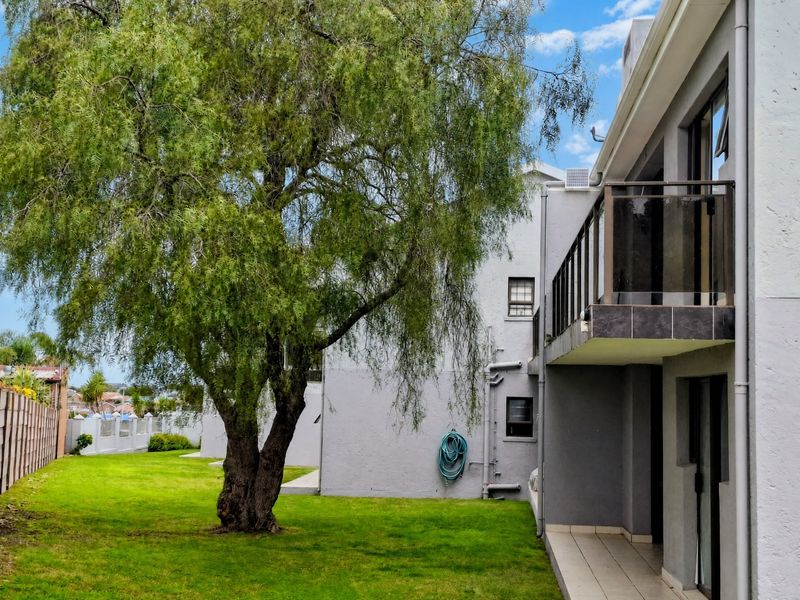 Rear view of the apartment building featuring a private balcony with glass balustrade, ground floor patio area, and access to a shared garden space.
