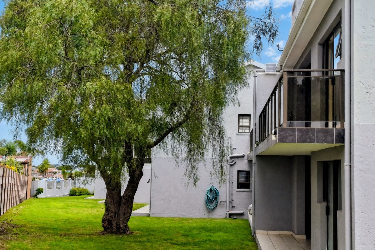 Rear view of the apartment building featuring a private balcony with glass balustrade, ground floor patio area, and access to a shared garden space.