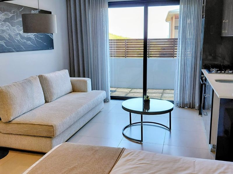 Modern open-plan living area featuring a compact kitchen with stone countertops, gas hob, and direct access to a private balcony through sliding glass doors.
