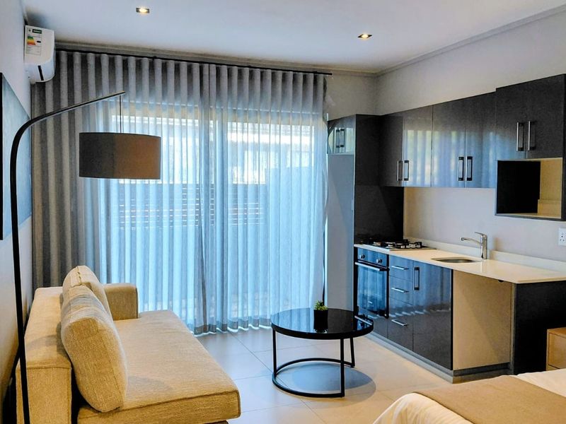 Modern open-plan living area featuring a compact kitchenette with dark cabinetry, integrated appliances, tiled flooring, and direct access to a balcony through sliding doors.