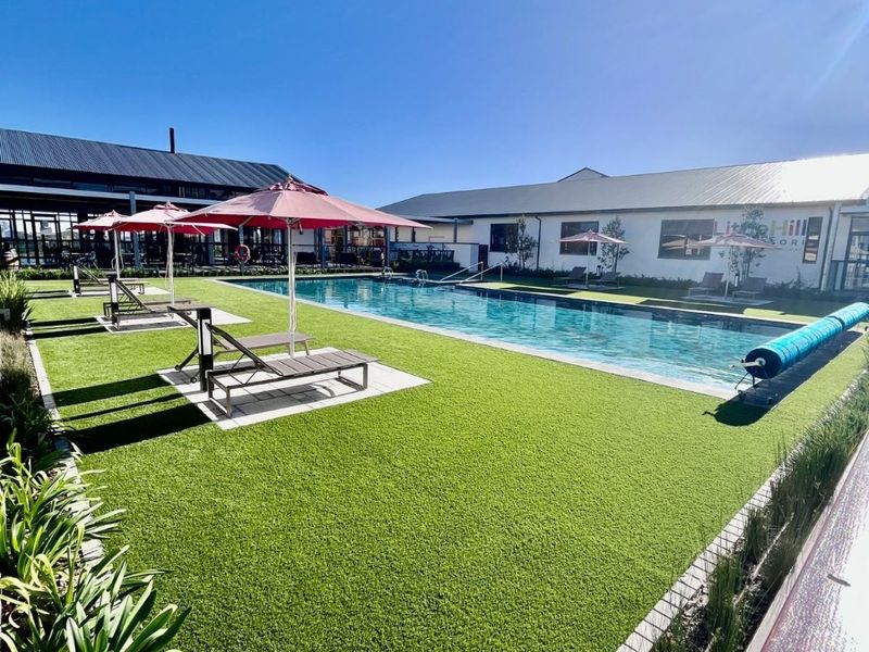 A well-maintained outdoor swimming pool area featuring a clear pool, artificial turf lawn, and paved surrounds, set within a resort-style environment.