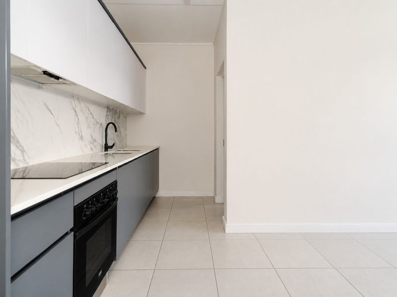 A modern, minimalist kitchen featuring sleek grey and white cabinetry, marble-look splashback, integrated electric hob, oven, and neutral tiled flooring.