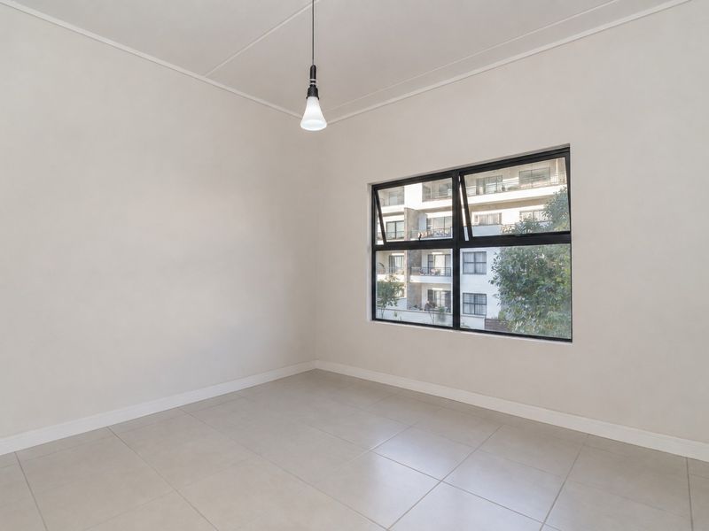 A bright, empty bedroom featuring neutral tiled flooring, clean white walls, and a modern black-framed window with a view of the surrounding apartment complex.