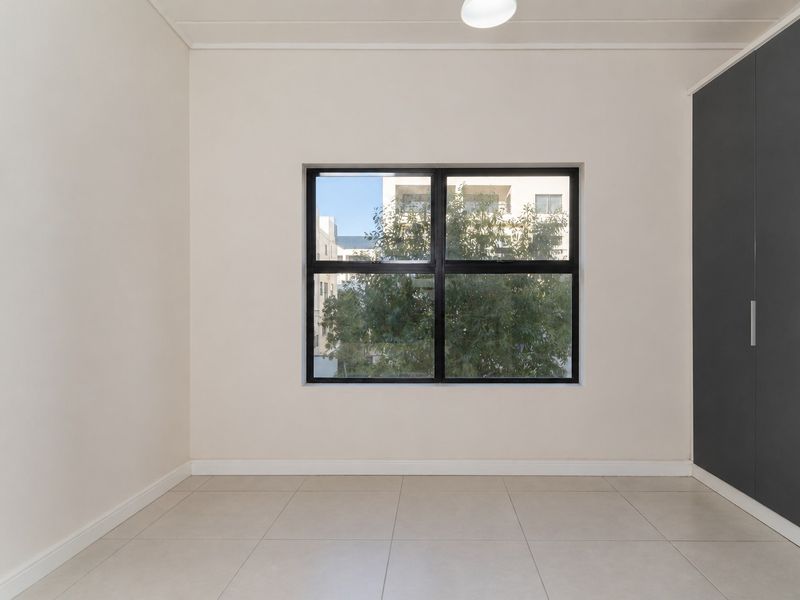 A bright, empty bedroom featuring neutral tiled flooring, a large black-framed window, and modern built-in cupboards.