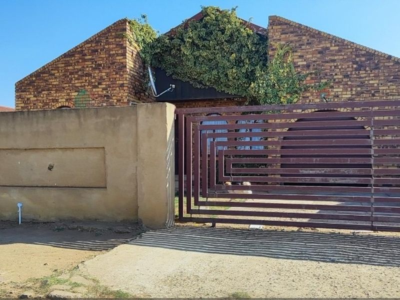 Front view of a face brick house featuring a metal sliding gate, perimeter wall, and driveway, with visible vegetation growth on the building facade.