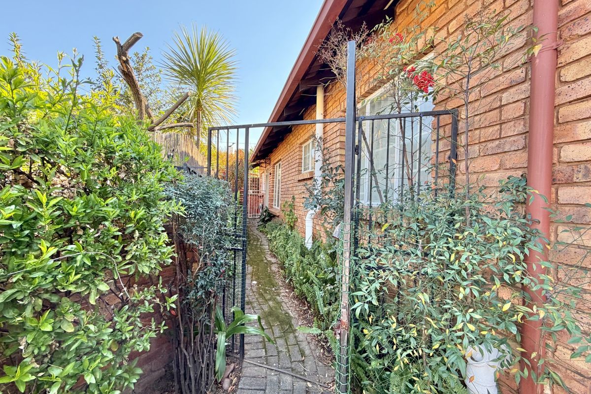 A side pathway featuring a brick facade, paved walkway, and metal security gate, surrounded by established garden greenery.