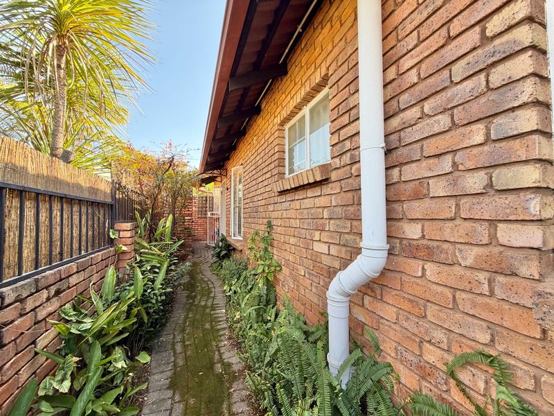 A side exterior view of the townhouse featuring a brick facade, a paved walkway, and established garden greenery along the boundary wall.