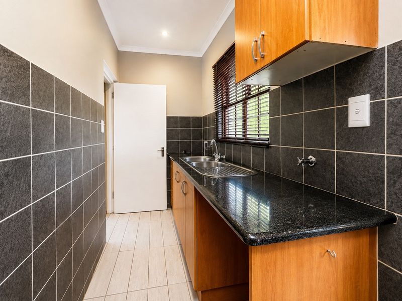 A functional scullery featuring granite countertops, double stainless steel sink, and ample wooden cabinetry with dark tiled splashbacks.