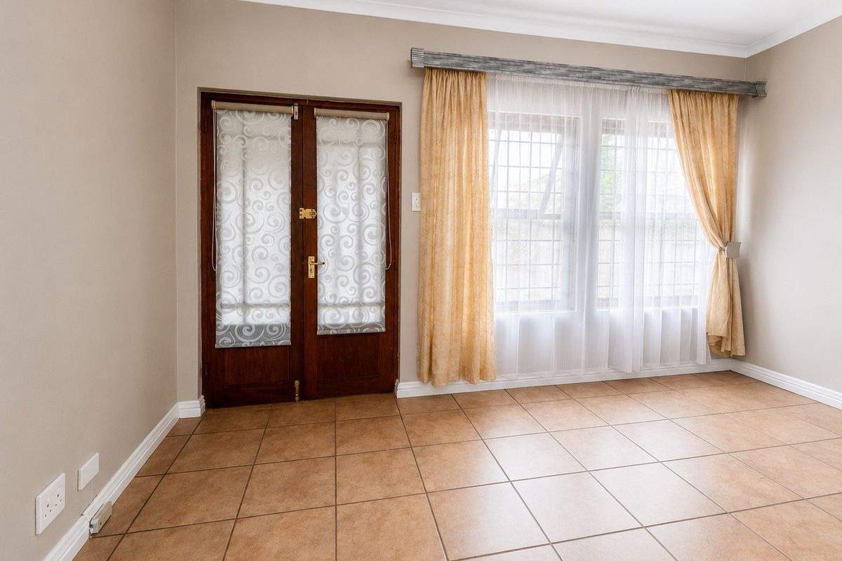 An empty living room featuring neutral walls, tiled flooring, double wooden doors, and large windows with security bars.
