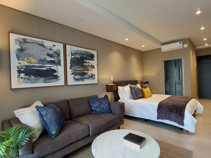 A modern open-plan studio apartment featuring neutral wall tones, recessed ceiling lighting, tiled flooring, and a wall-mounted air conditioning unit.