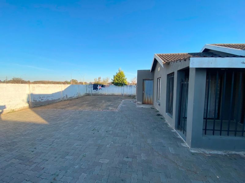 Rear exterior view of the property featuring a paved courtyard, perimeter wall, and a single-story structure with security bars on windows.
