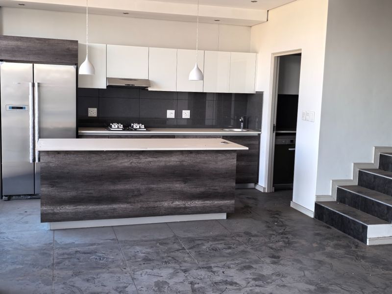 Modern open-plan kitchen featuring dark wood cabinetry, a central island, and a sleek staircase with a glass balustrade in a penthouse setting.