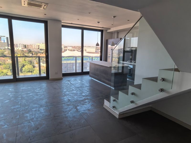 Spacious open-plan living and kitchen area featuring a modern staircase with glass balustrades, large floor-to-ceiling windows, and access to a balcony with city views.