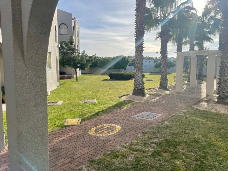 A well-maintained communal garden area featuring a paved walkway, established palm trees, and a pergola structure within an apartment complex setting.