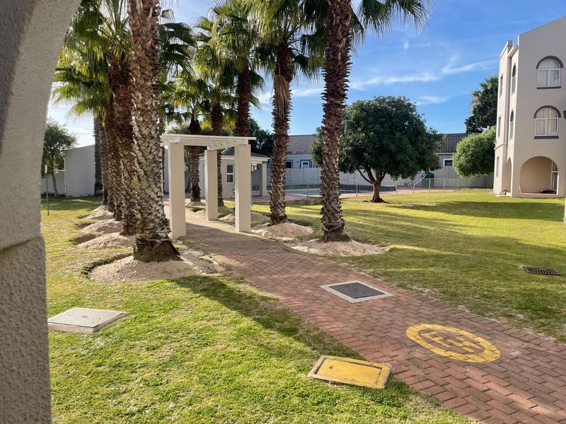 A well-maintained communal garden area featuring a paved walkway, mature palm trees, and a pergola, providing a pleasant outdoor space for residents.