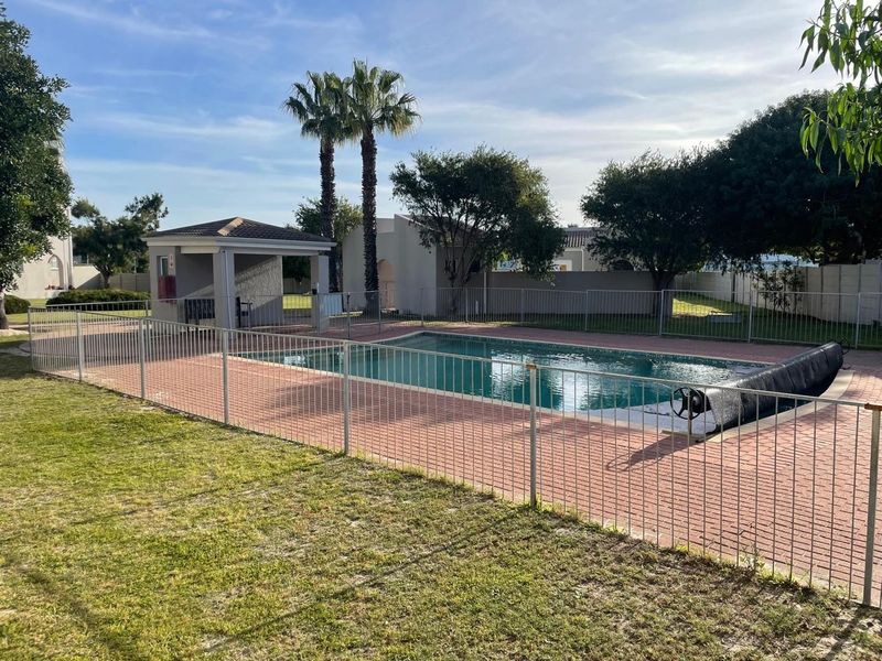 Communal swimming pool area featuring a paved deck, safety fencing, and a covered gazebo structure within a landscaped garden setting.