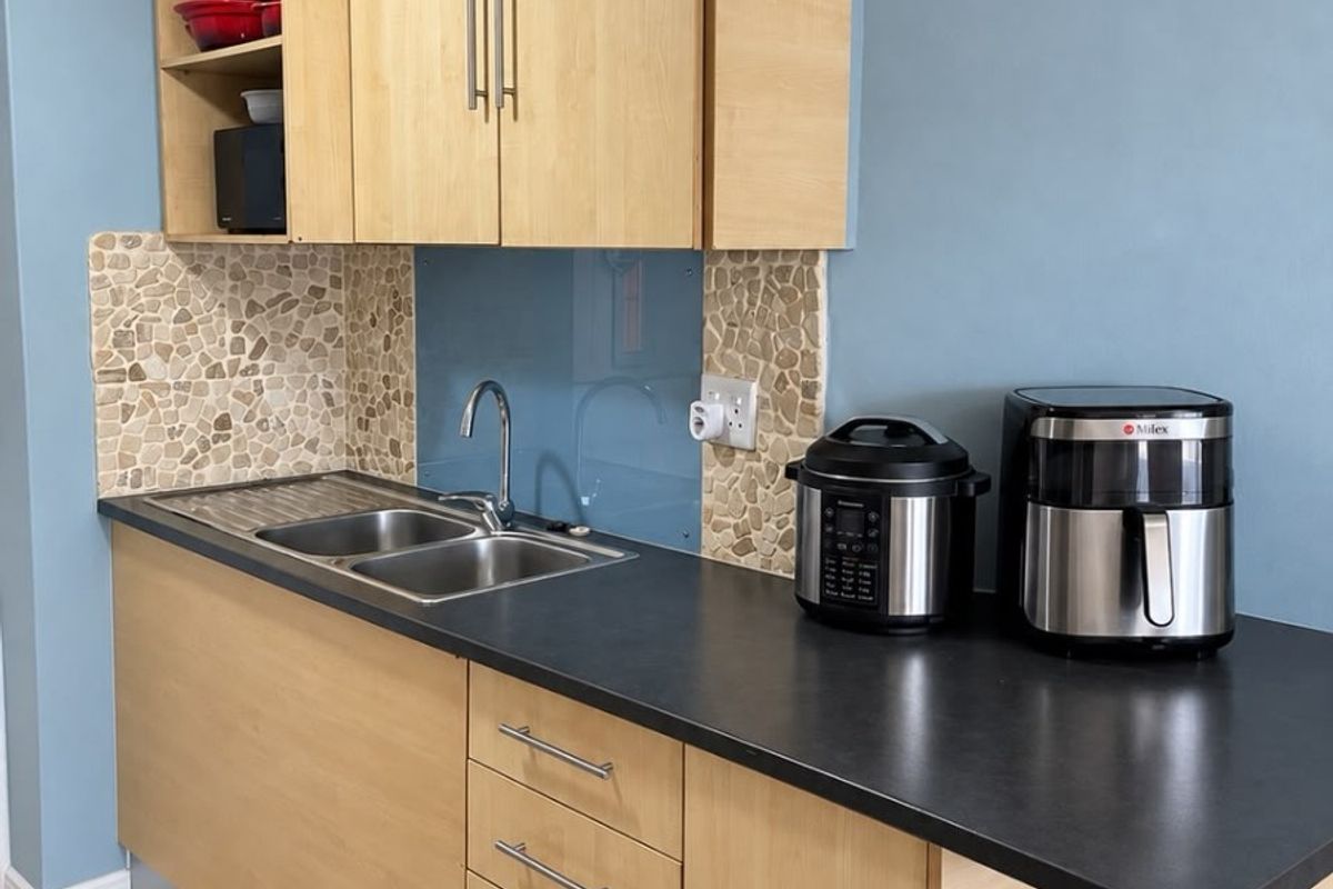 A compact kitchen featuring light wood-tone cabinetry, a dark laminate countertop, stainless steel sink, and a combination of stone mosaic and glass splashbacks.