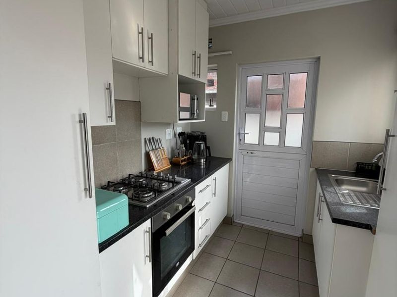 A functional galley-style kitchen featuring white cabinetry, dark granite countertops, a gas hob, built-in oven, and tiled flooring with direct exterior access.