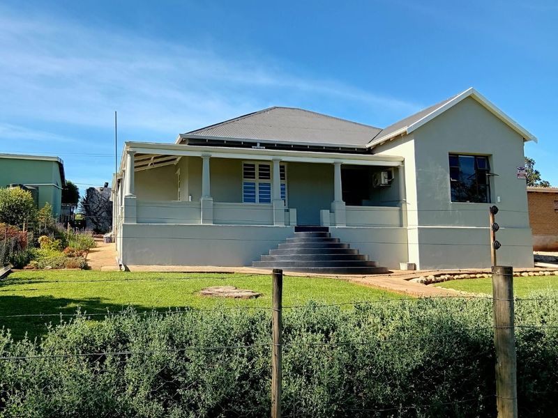 A well-maintained single-story house featuring a covered front veranda, concrete entrance stairs, and a neat garden with perimeter electric fencing.