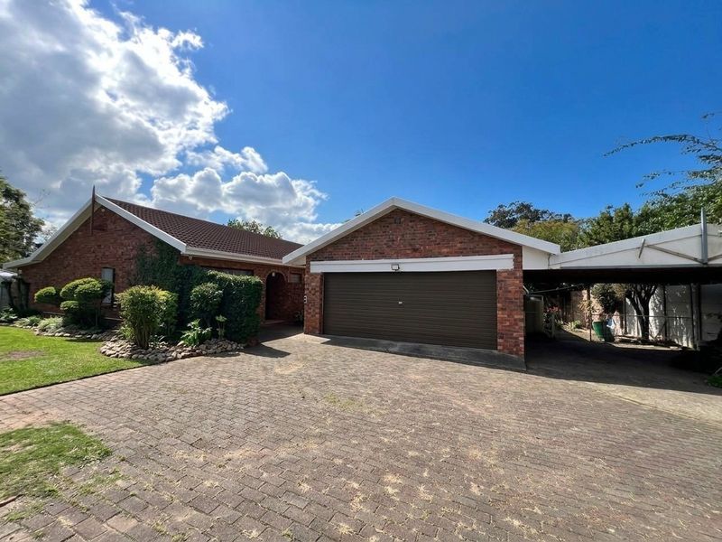 Front exterior view of a brick house featuring a paved driveway, a garage, and an attached carport with a landscaped garden area.