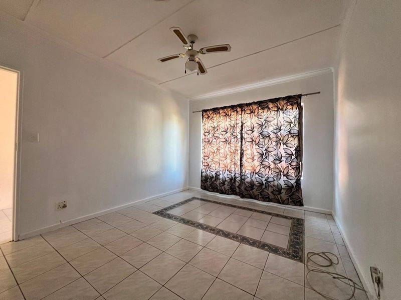 A bright, empty living room featuring neutral tiled flooring with a decorative border and a ceiling fan.