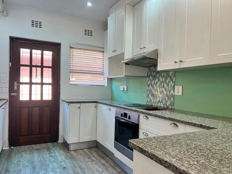 A clean, functional kitchen featuring white cabinetry, granite countertops, built-in oven, and a mosaic tile backsplash with direct access to the exterior.