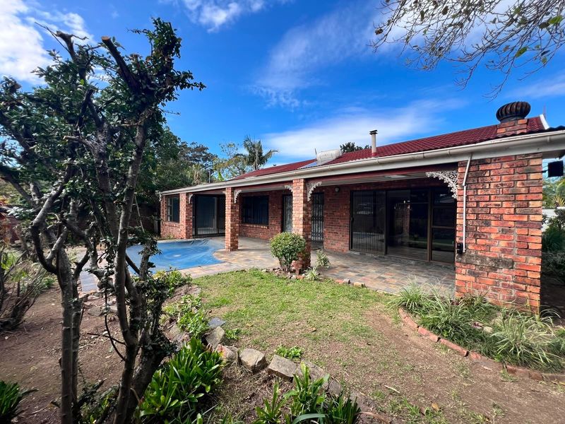 Rear view of a brick house featuring a covered patio, swimming pool, and established garden area under a tiled roof.