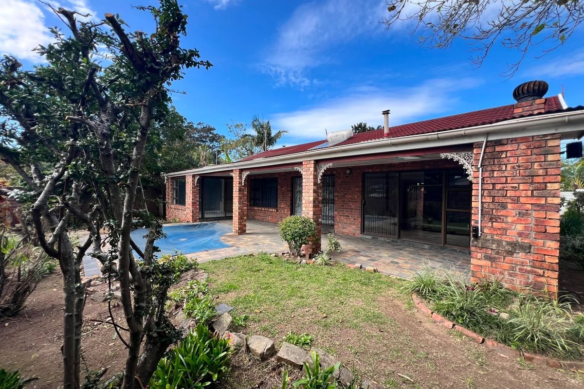 Rear view of a brick house featuring a covered patio, swimming pool, and established garden area under a tiled roof.