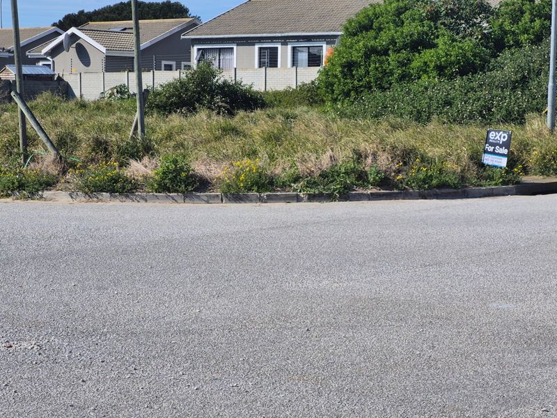 Street view of a vacant residential plot featuring asphalt road frontage and concrete kerbing, with a real estate for sale sign visible.