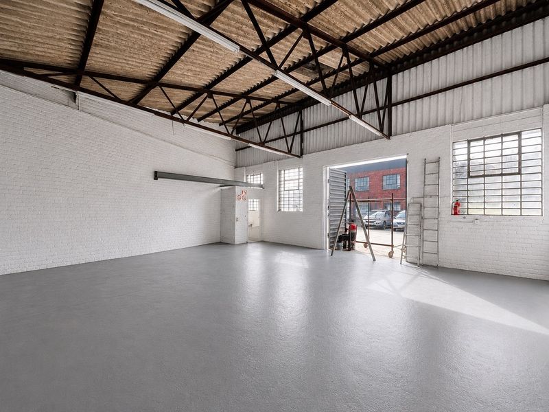Spacious industrial warehouse unit featuring high ceilings with exposed steel trusses, painted brick walls, durable epoxy flooring, and large roller shutter access.