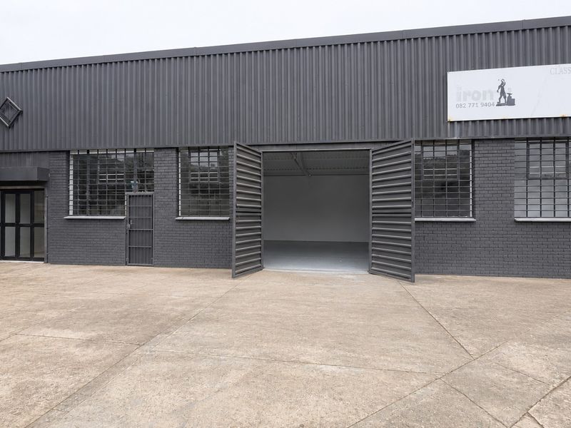 Industrial warehouse exterior featuring dark grey corrugated metal cladding, brick facade, secure window bars, and a spacious concrete forecourt with large swing door access.