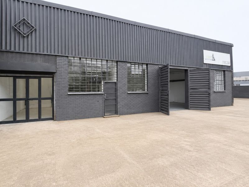 Industrial property exterior featuring dark painted brickwork, corrugated metal cladding, large swing doors, and a spacious concrete forecourt area.