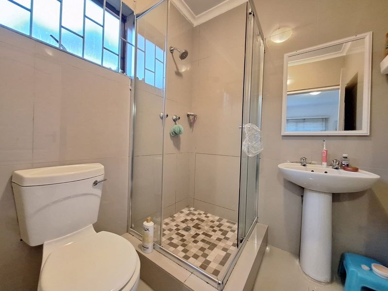 A functional bathroom featuring a pedestal basin, a glass-enclosed shower, and a standard toilet with neutral wall and floor tiling.