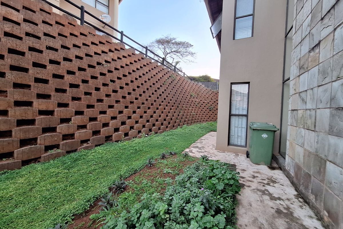 Rear exterior view of the property featuring a large tiered retaining wall, stone-clad facade, and a landscaped garden area with a paved walkway.
