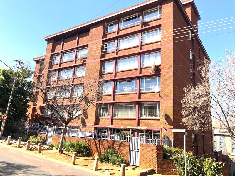 A multi-story apartment building featuring a red brick facade, security fencing, and gated access from the street.