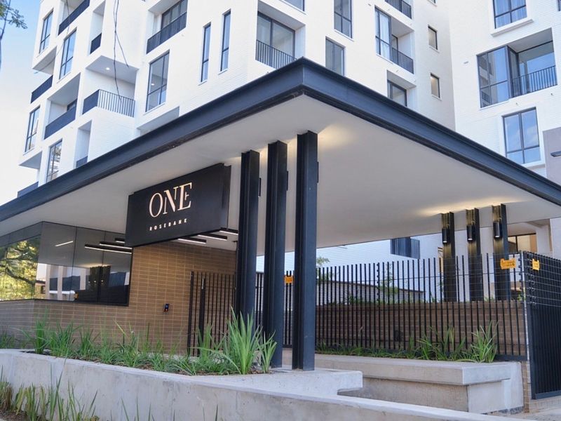 Modern apartment building entrance featuring a sleek covered canopy, steel structural columns, and secure perimeter fencing with integrated lighting.