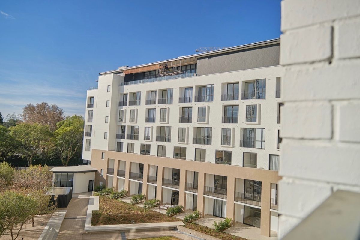 A view of the rear facade of a modern apartment building featuring multiple balconies, glass balustrades, and a landscaped garden area.