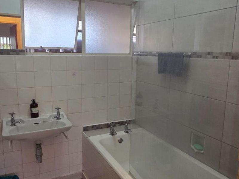 A functional bathroom featuring a wall-mounted basin, a bathtub with a glass shower screen, and neutral wall and floor tiling.