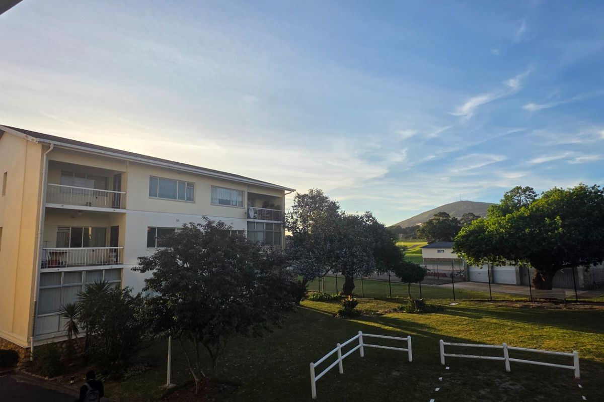 Exterior view of a multi-story apartment building featuring balconies, surrounding garden space, and a paved driveway area under a clear sky.