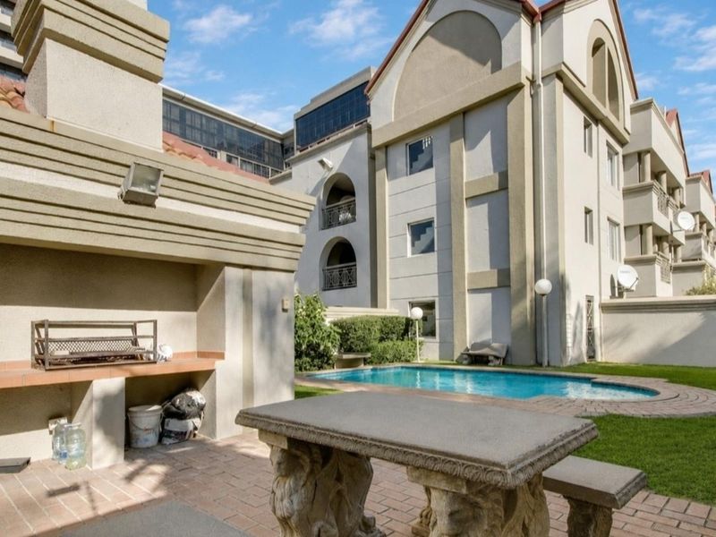 Communal outdoor area featuring a swimming pool, paved patio, and a built-in braai facility within a well-maintained apartment complex.