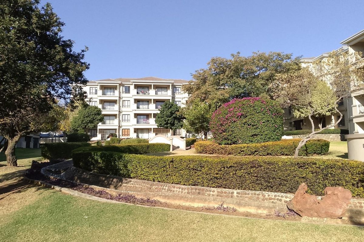 A well-maintained apartment complex exterior featuring landscaped gardens, manicured hedges, and a stone retaining wall leading to the building facade.
