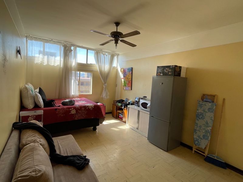 Open-plan studio apartment featuring a ceiling fan, large windows for natural light, and vinyl flooring, offering a functional living and sleeping space.