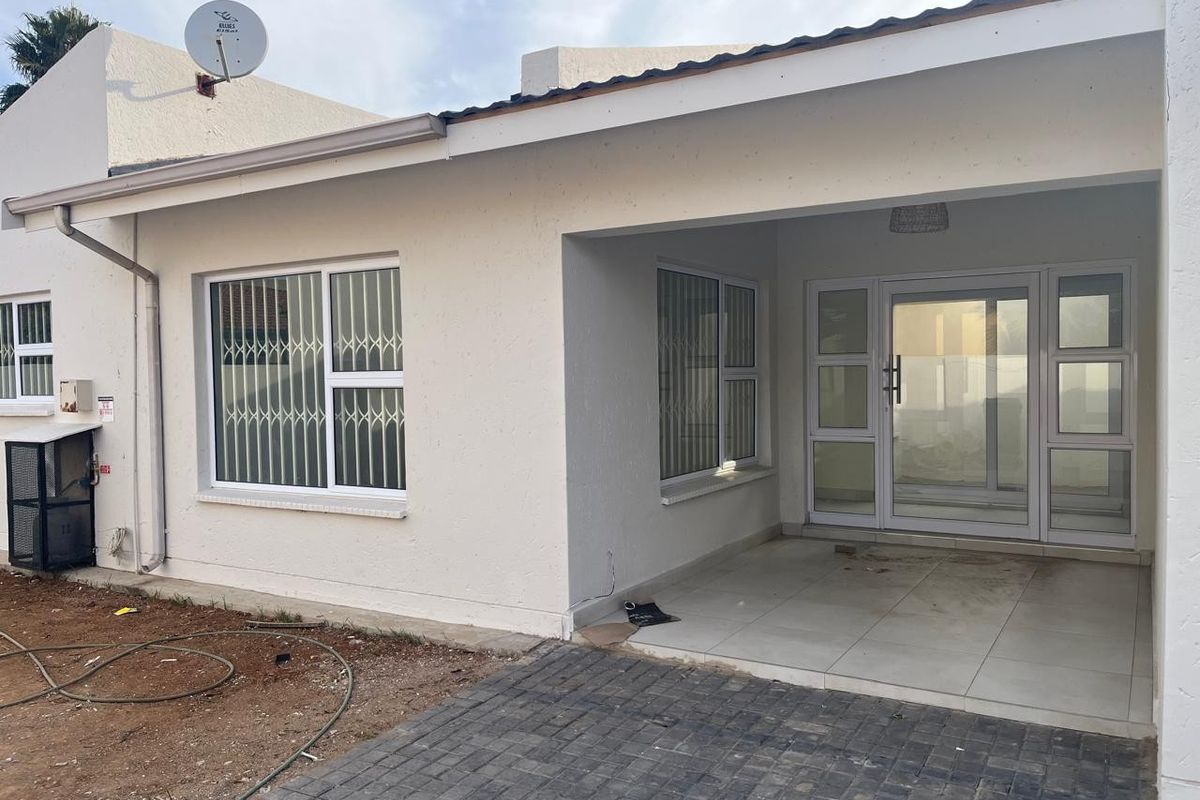 Rear exterior view featuring a covered tiled patio, aluminium sliding doors, and security-fitted windows, leading onto a paved area with unfinished garden space.